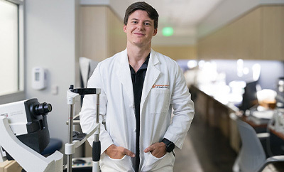 Kirby Taylor wears a medical white coat and stands for a portrait in an eye exam room.