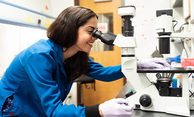 Inside a research lab, a woman wearing a blue research lab coat looks into a microscope while smiling.